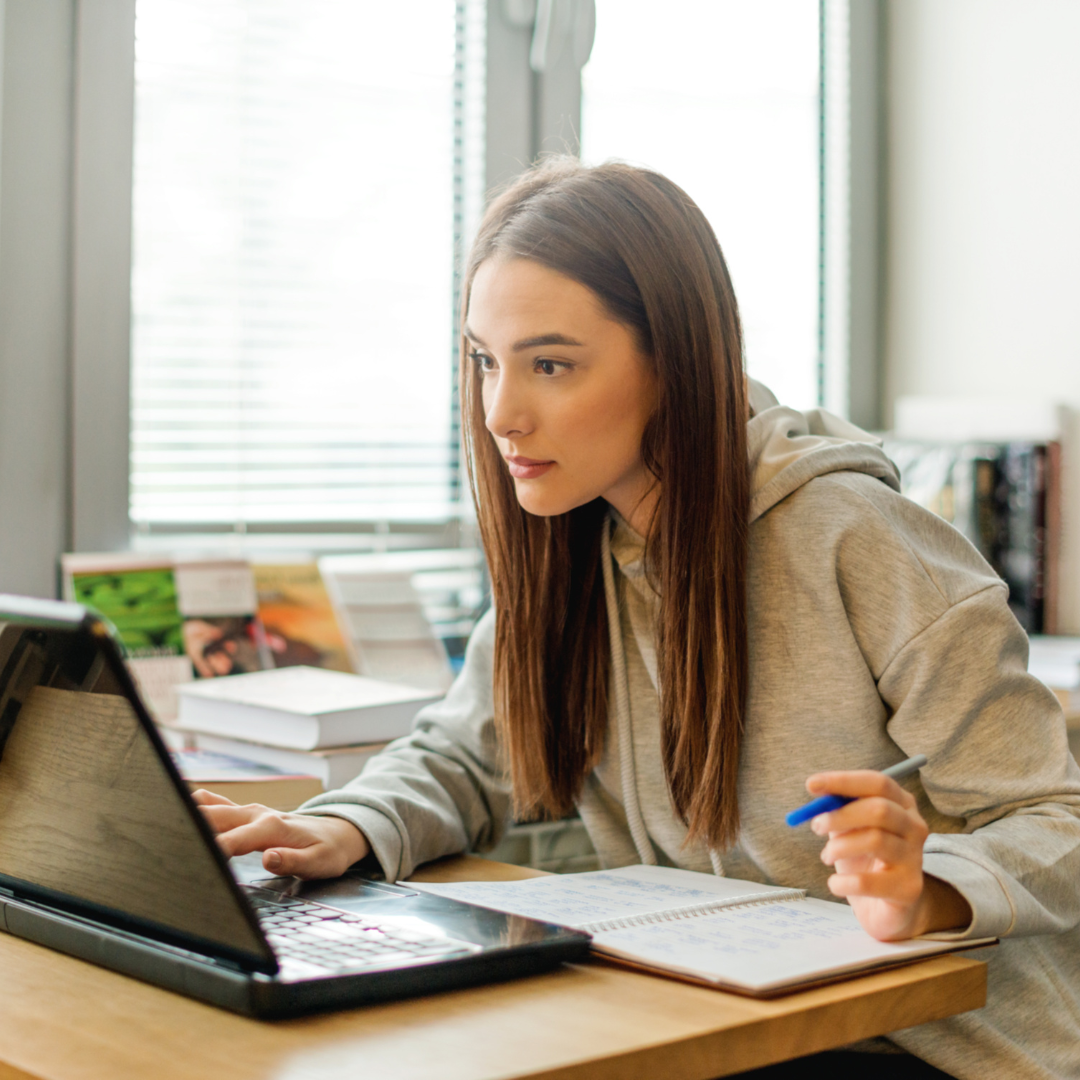 Woman sitting at a desk with a computer and notebook, her left hand has a pen and right hand on the keyboard