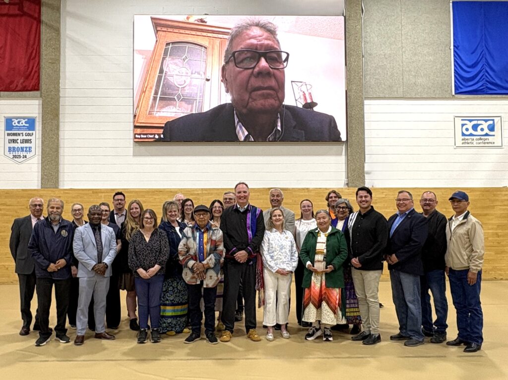 StevefullgroupRoy2 - Portage College Group of diverse people posing together in a gymnasium, with a large video screen showing a man in glasses in the background.