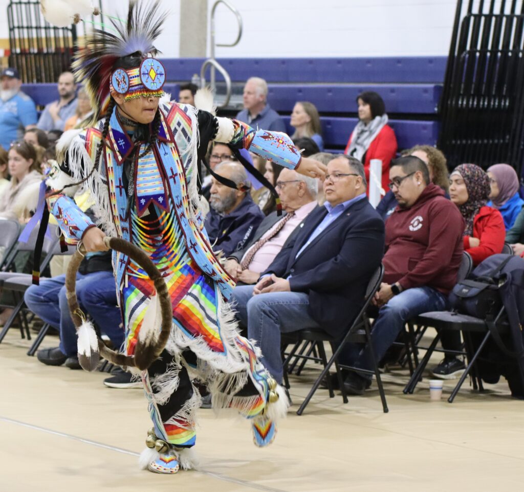 sdancerGaryLameman1 - Portage College Dancer in bright, beadwork regalia and feather headdress performing a powwow-style dance in a gymnasium with seated spectators in the background.