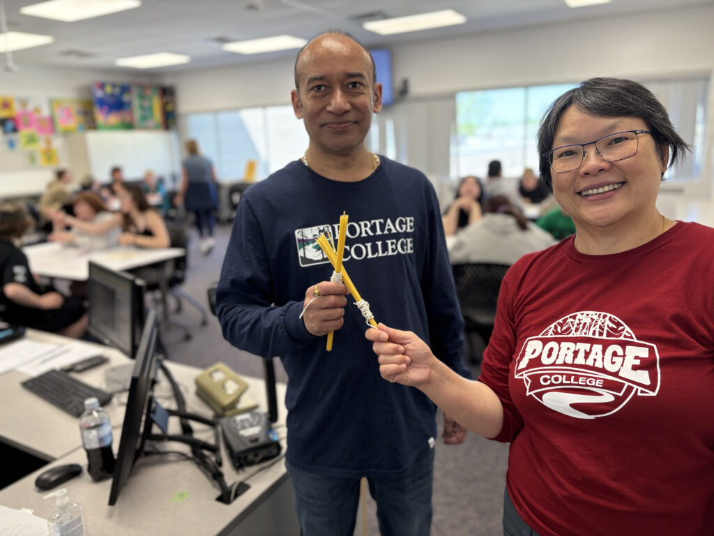 vinodmerlynbusiness - Portage College Two adults smiling for a selfie in a busy classroom; man wears a Portage College shirt and holds yellow cords, woman beside him wears a red Portage College shirt.