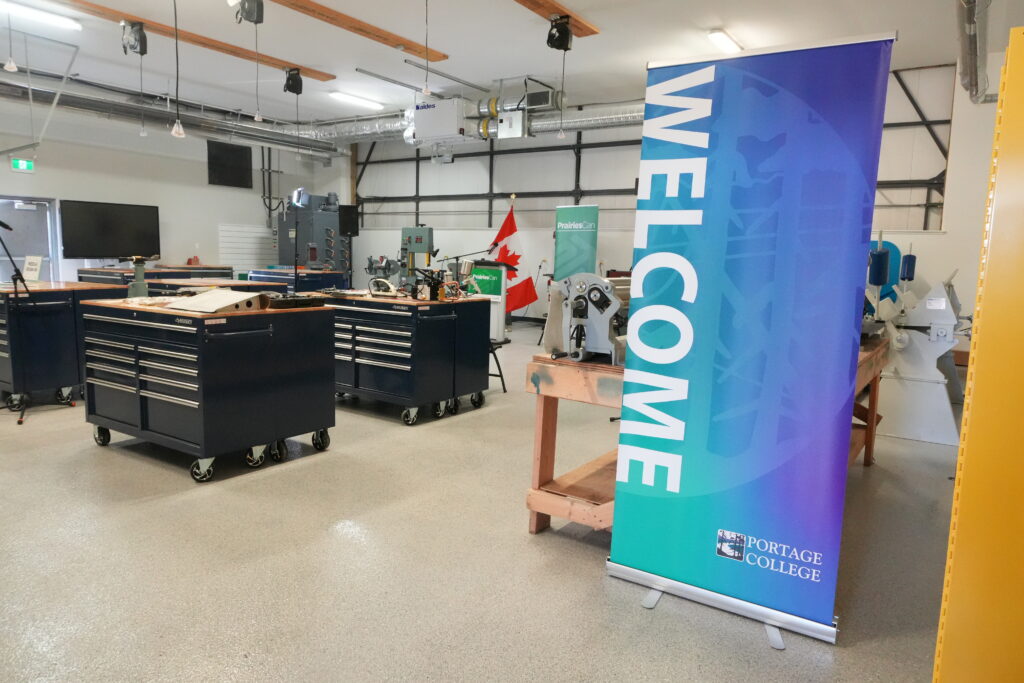 1DSC01224 - Portage College Spacious workshop with dark tool chests, workbenches, and a tall blue gradient banner in the foreground; Portage College logo at the bottom edge.