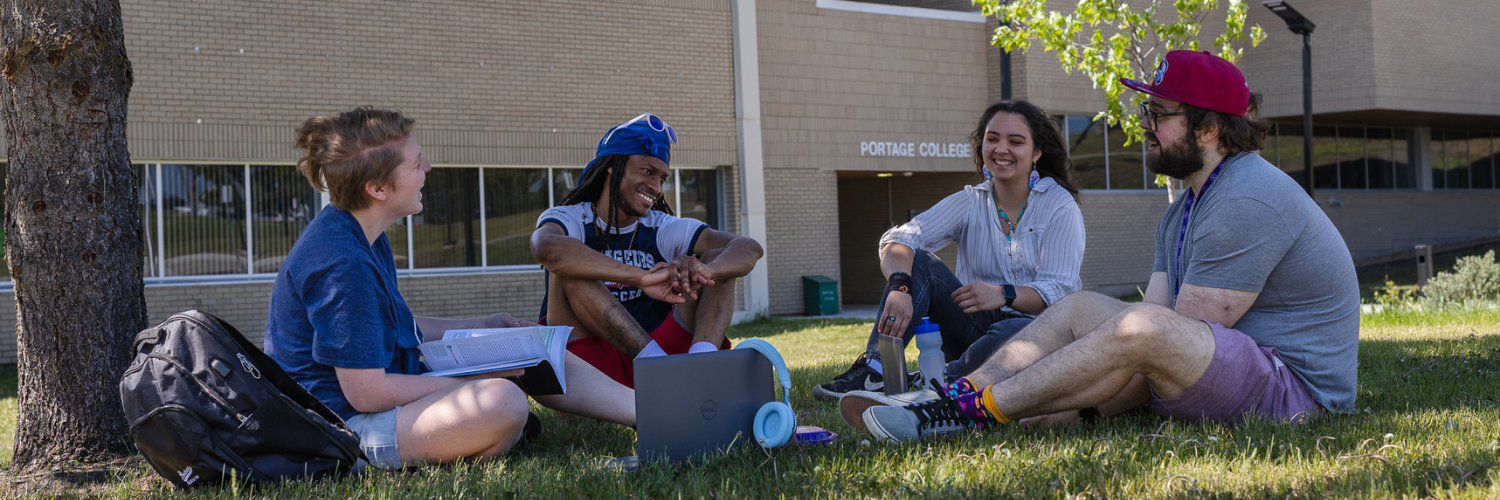 Four students sit on the campus lawn outside a brick building, studying and chatting with a laptop and books nearby.