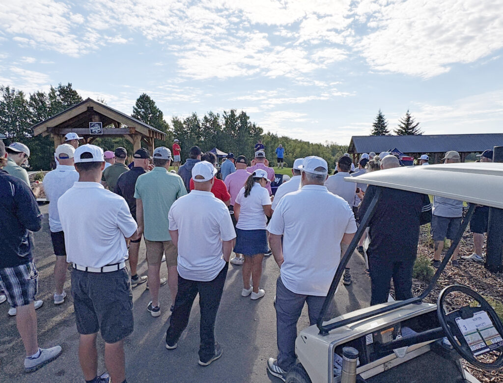 Crowd of people outdoors at an event, many wearing white baseball caps and casual clothing, facing a small stage or speaker area.
