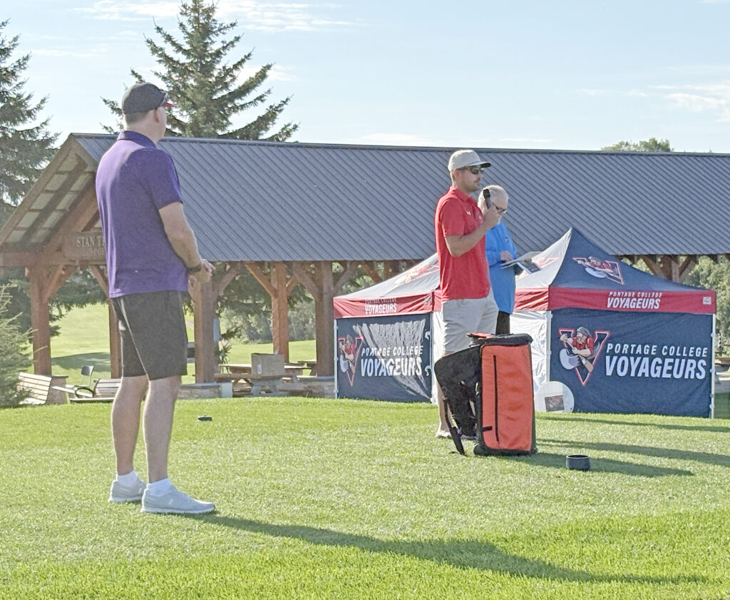 Man in a purple shirt facing away stands on a grassy field beside a Portage College Voyageurs tent, with a man in red speaking into a microphone nearby.