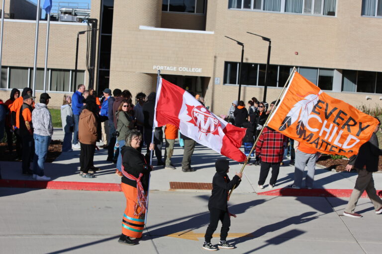 People gather outside Portage College, waving red-and-white and orange banners on a sunny day.