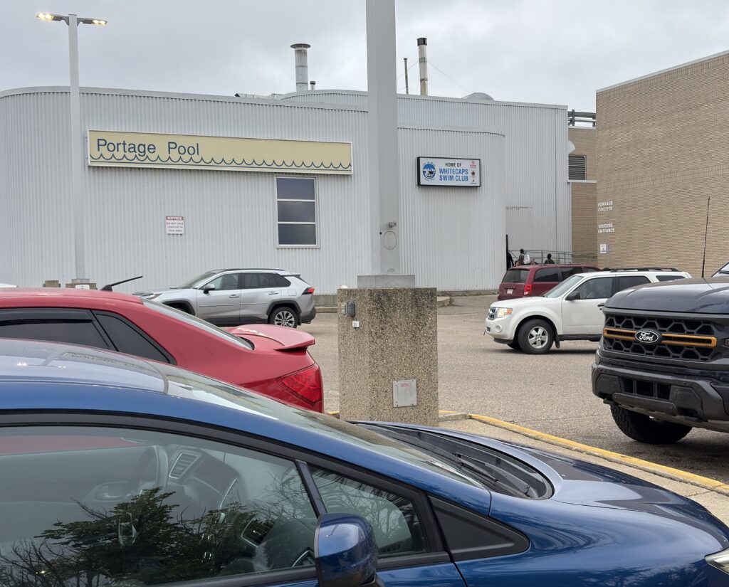 Parking lot in front of a white industrial building with a Portage Pool sign and a Whitecaps Swim Club sign on the wall, several cars visible.