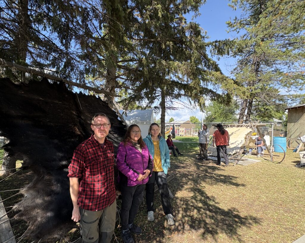 IMG_8988 1 - Portage College People standing in a line beside a large black animal outdoors, with trees overhead and a chain-link fence in the background.