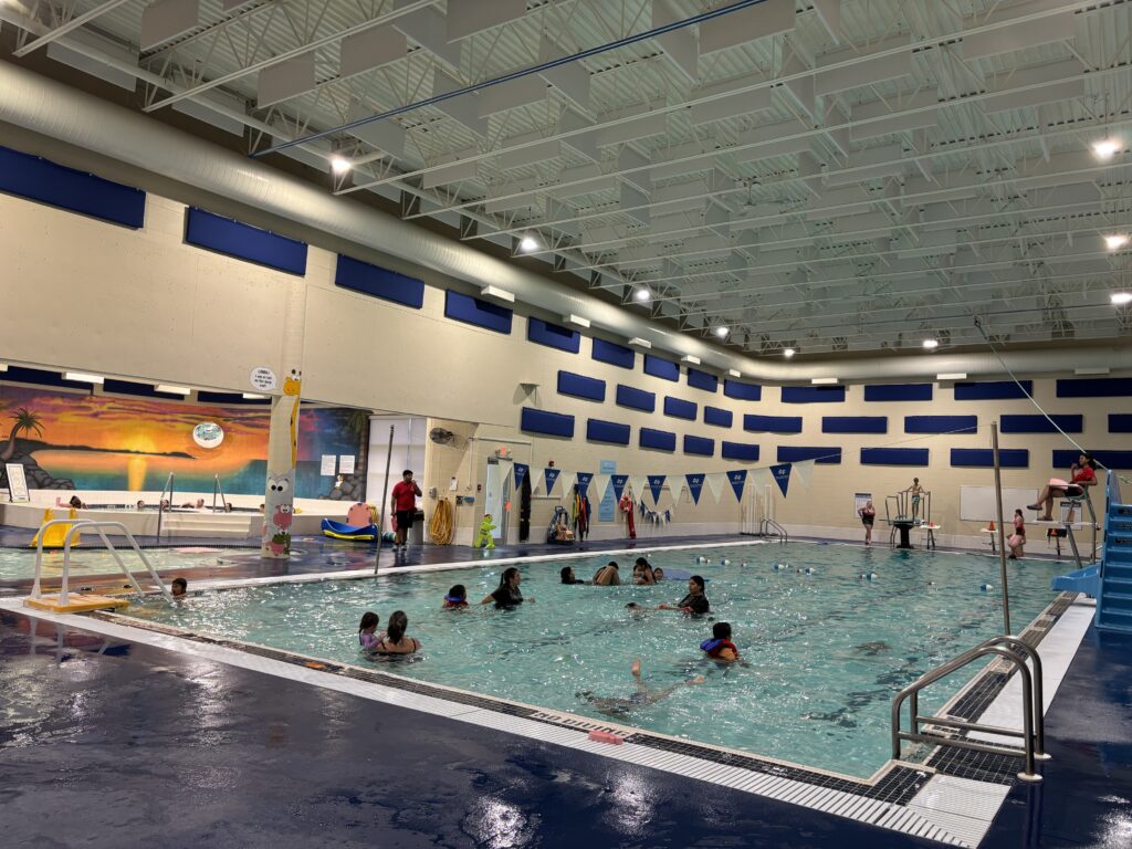 Indoor swimming pool area with swimmers and a tropical beach mural on the far wall, blue wall pads along beige walls, and a white ceiling structure.