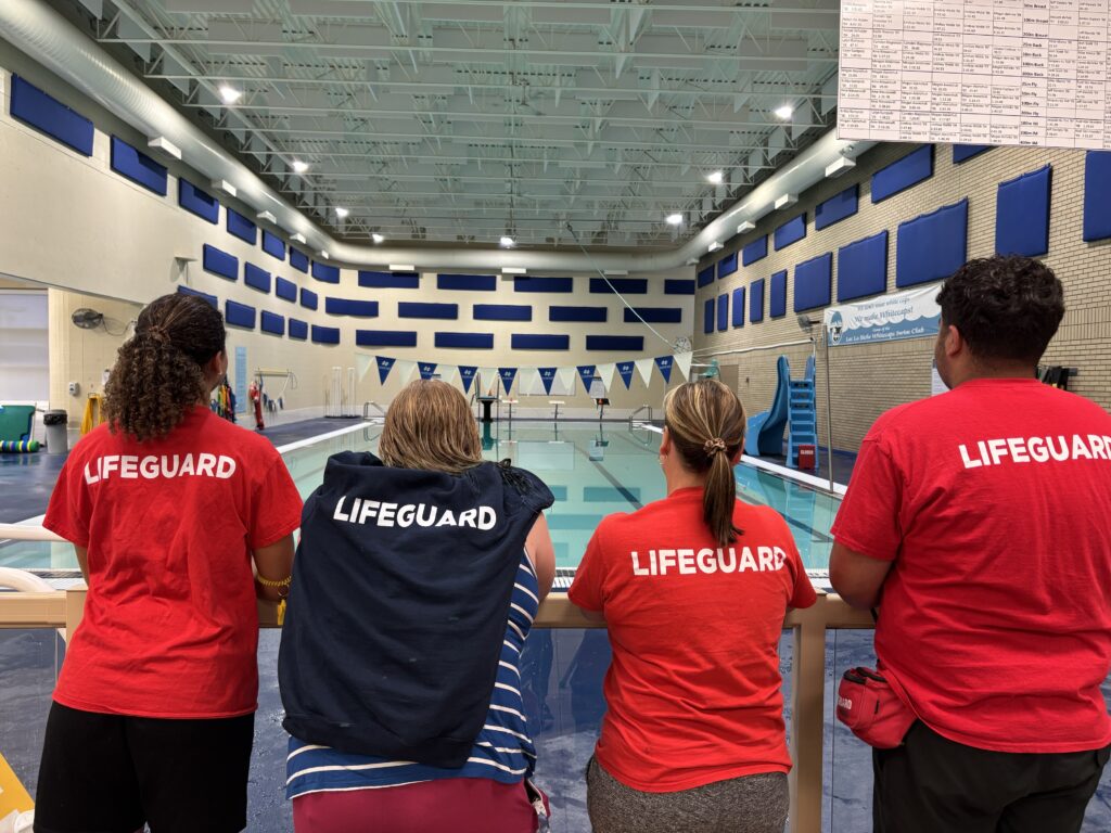 Team of lifeguards standing at poolside, backs to camera, wearing red shirts with LIFEGUARD printed on back.