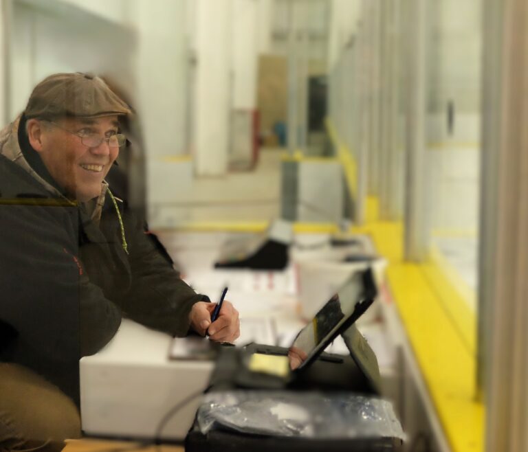 Smiling elderly man wearing a brown cap and glasses, signing on a tablet at a checkout counter.