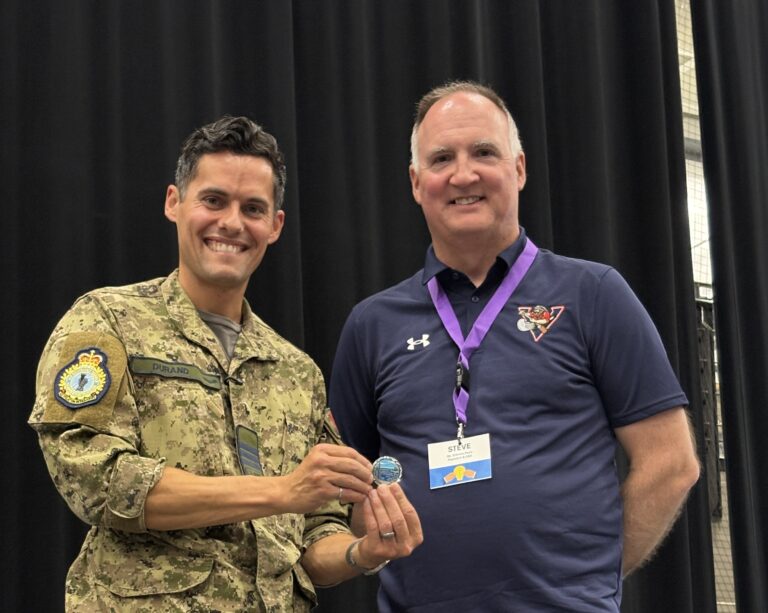 Two men posing and smiling in front of a black curtain; one in camouflage uniform with a patch, the other in a navy polo with a purple lanyard nearby.