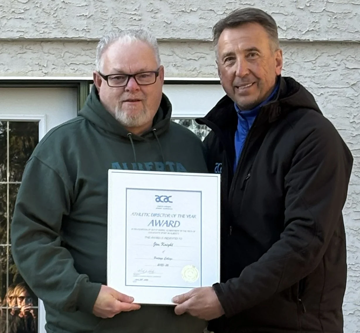Two men stand outside a house, one presenting a framed certificate to the other.