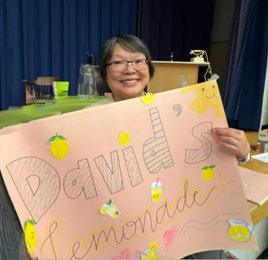 MerlynwithSign copy - Portage College Smiling woman holding a large pink poster that reads'David's memories' with lemon drawings and lemonade icons on a stage.