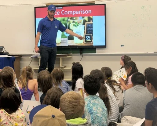 davidwindow2 - Portage College Instructor in a blue polo and cap points to a large screen that reads'Price Comparison' as students sit on the floor watching in a classroom.