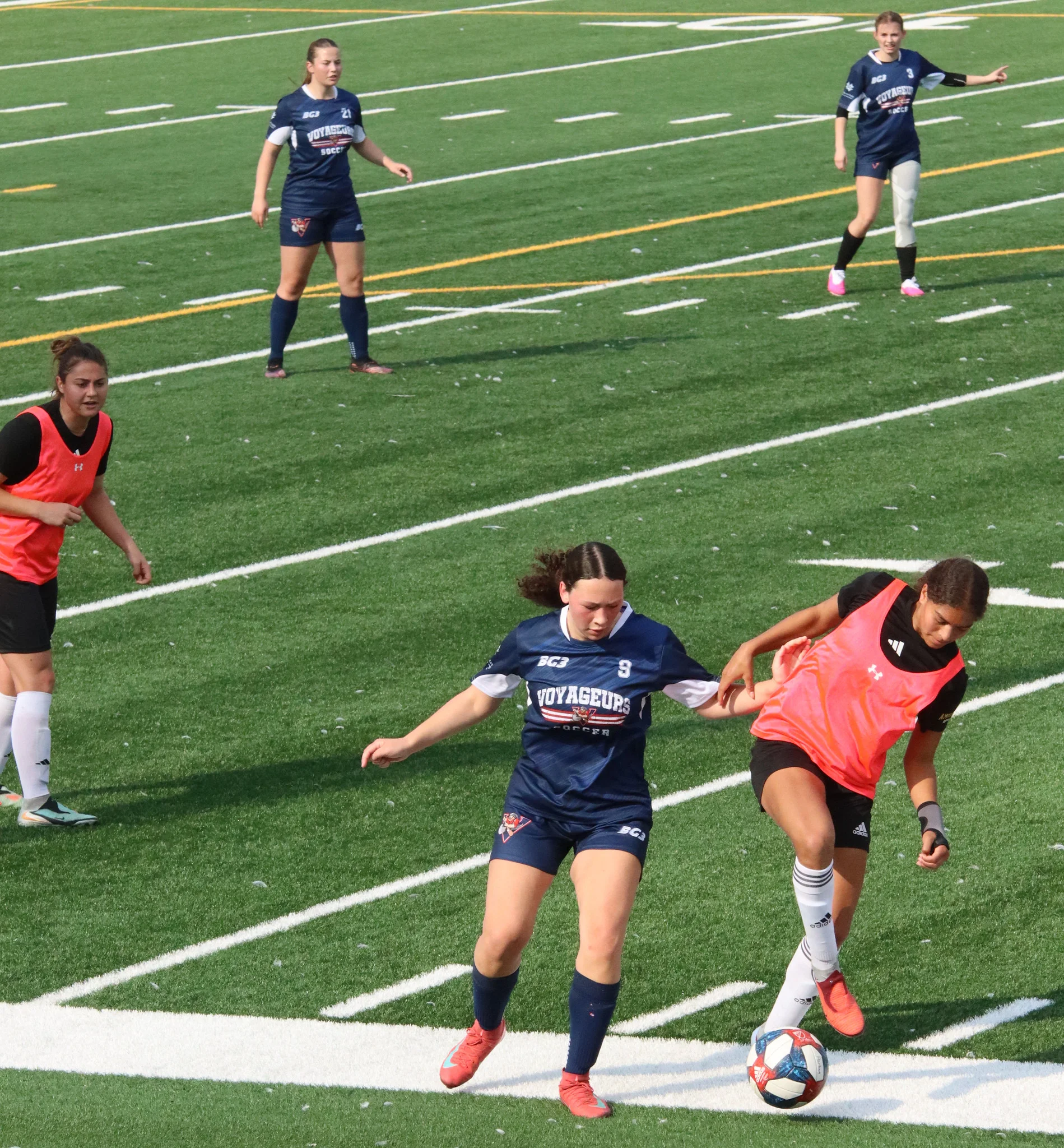 Girls' soccer match on artificial turf; a player in navy with number 9 reaches for a ball near the sideline as teammates in blue and pink bibs position nearby.