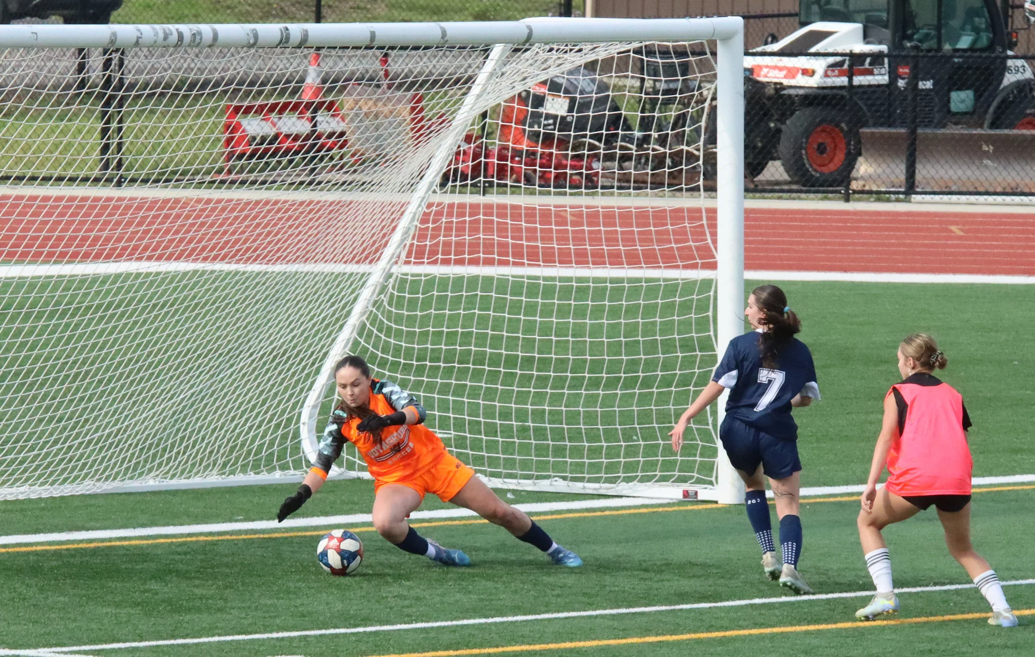 Soccer goalkeeper in orange dives toward the ball near the goal during a match on artificial turf.