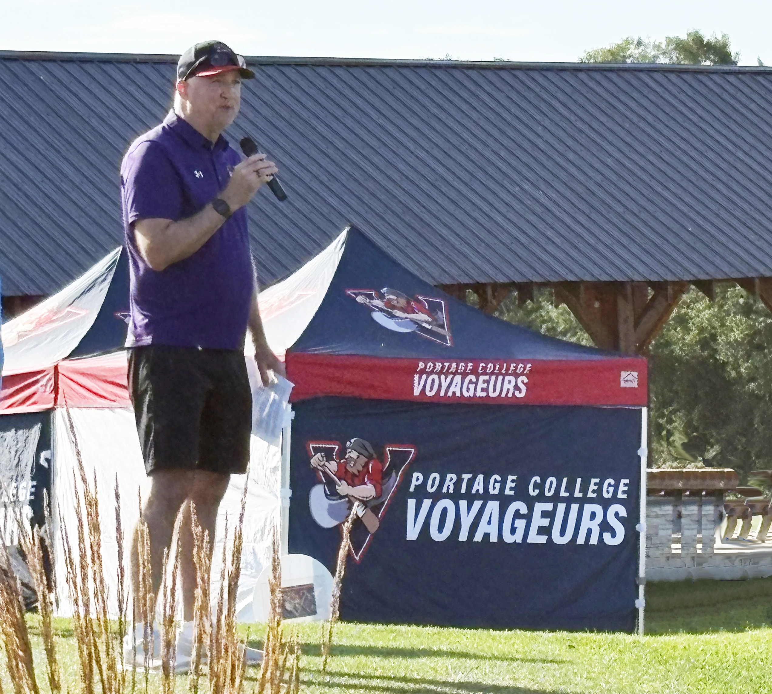 Man in a purple polo speaking into a handheld microphone on an outdoor stage with Portage College Voyageurs banners behind him.