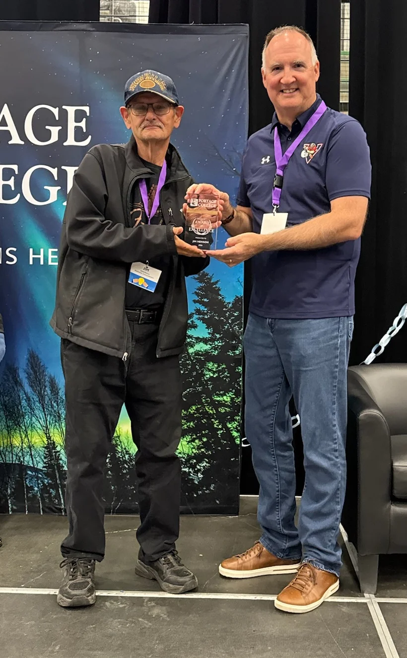 Two men on a stage exchange a glass trophy during an award ceremony; the man on the right wears a purple lanyard and navy polo, the other in a black jacket and cap.