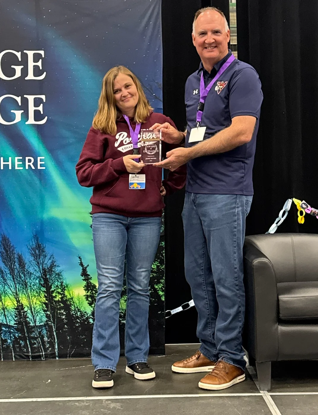 A woman in a maroon hoodie accepts an award from a man in a navy polo on a stage, with a night-sky backdrop and trees.