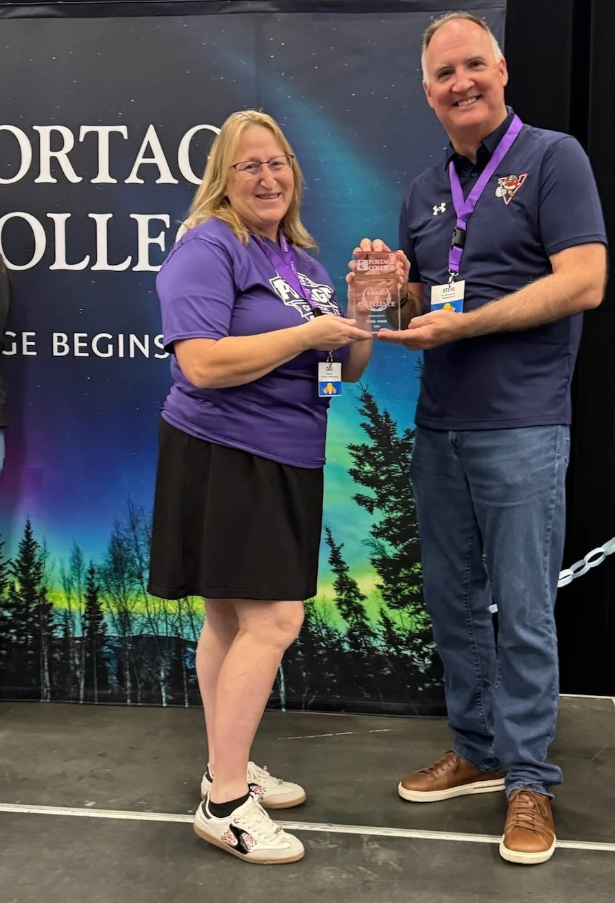 Two adults on a stage exchange a glass award, smiling, with a forest- and night-sky backdrop behind them.