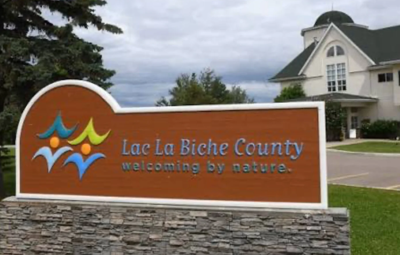Lac La Biche County welcome sign with colorful logo and the phrase'welcoming by nature' in front of a stone base and a building nearby.