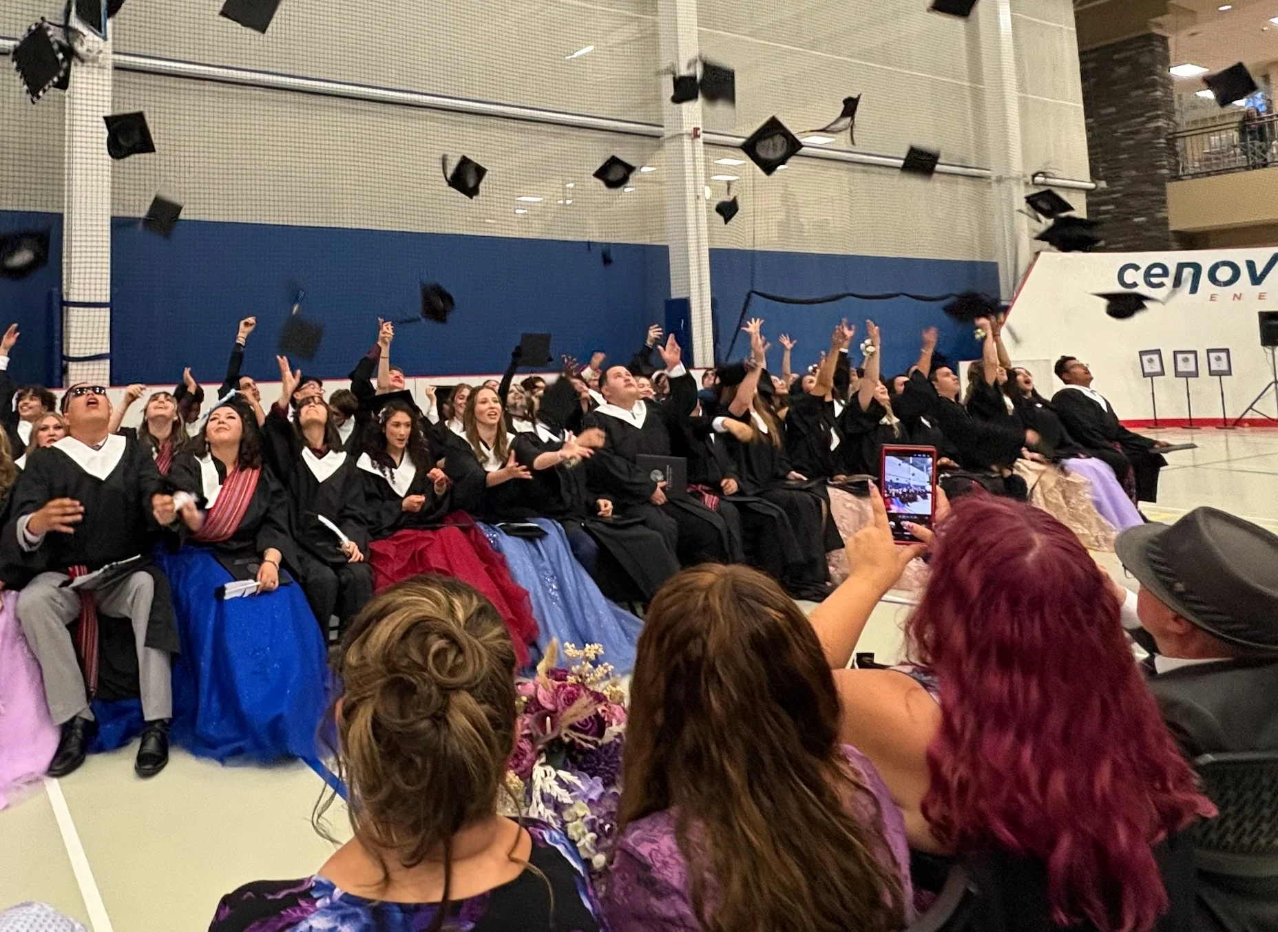 Group of graduates in caps and gowns throwing caps in the air during a ceremony in a gymnasium, with an audience cheering and taking photos nearby.